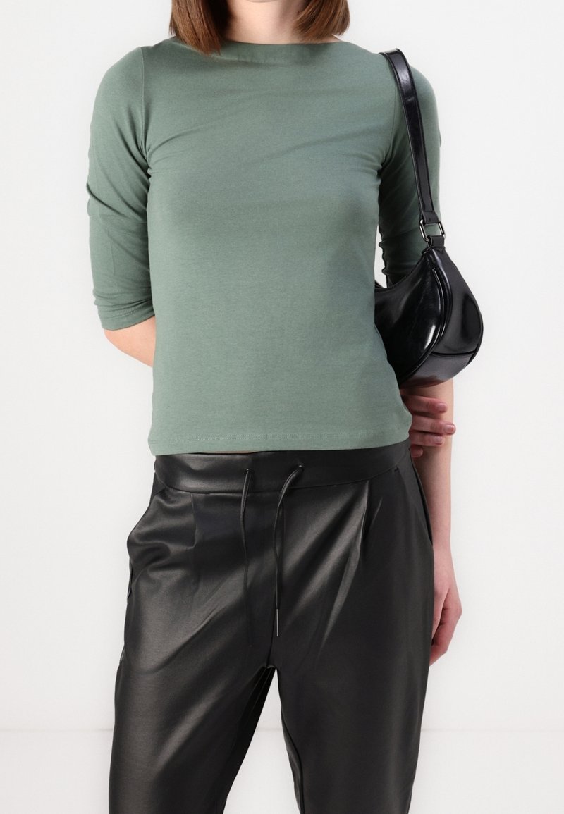 Femme portant un haut vert à manches longues, un pantalon en cuir noir avec cordon de serrage et un sac à épaule noir sur fond blanc.