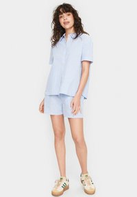 Camicetta azzurro chiaro e bianca a righe con maniche corte, abbinata a pantaloni corti coordinati, indossata con sneakers color crema con accenti verdi.