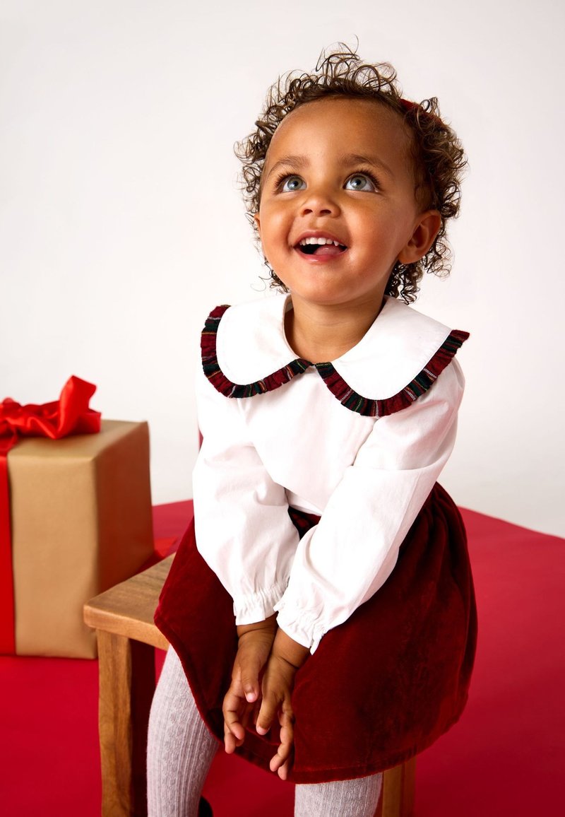Copil mic cu păr creț, purtând o bluză albă și o fustă de catifea roșie, așezat pe un scaun de lemn, privind în sus și zâmbind, cutie cadou cu panglică roșie în apropiere.