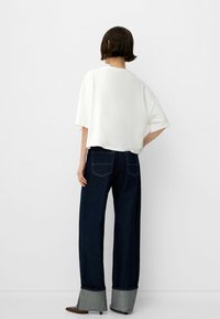 Camiseta corta blanca de gran tamaño combinada con jeans de pierna ancha azul oscuro que cuentan con puños de mezclilla gris claro y costuras en contraste.