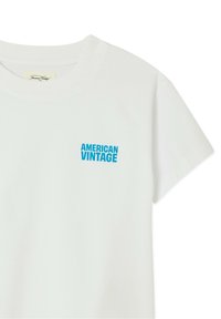 Biele tričko s krátkym rukávom s modrým nápisom "AMERICAN VINTAGE" potlačeným na ľavej hornej časti hrudníka.