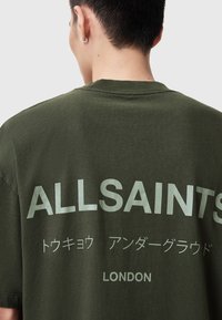 Camiseta de gran tamaño en verde oliva con cuello redondo, que presenta un gran texto "ALLSAINTS" en verde claro en la parte posterior, junto con caracteres japoneses y "LONDRES."