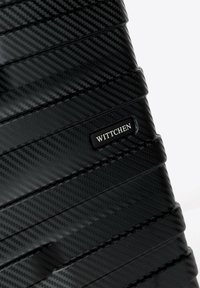 Černý tvrdý kufr s texturovaným vzorem carbonových vláken a horizontálními drážkami. Obsahuje vyraženou logo destičku s nápisem "WITTCHEN."