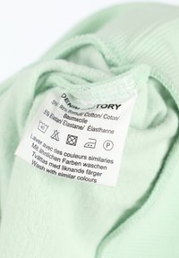 Tessuto verde menta con una consistenza morbida presenta un'etichetta di cura. L'etichetta elenca il materiale (95% cotone, 5% elastan) e le istruzioni per la cura in diverse lingue.