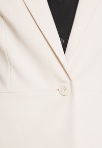 Blazer blanc au design épuré, texture lisse et fermeture à gros bouton blanc à l'avant. En arrière-plan, une chemise noire.
