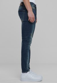 Tumedat sinised skinny teksad, millel on kulunud tekstuur ja minimaalne kulumus reitel, koos valgete tossudega.