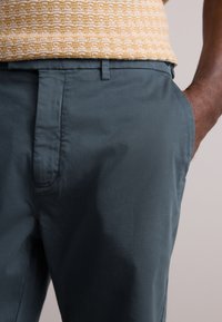 Tamsiai žalių petrolio spalvos kelnės pasižymi lygiu tekstūra, plokščiu priekiu ir šoninėmis kišenėmis, derinamos su raštuotu geltonu viršumi. Matomi subtilūs siuvimo detalės.