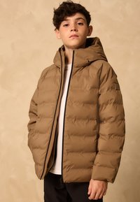 Khaki värvi padi-tüüpi jope kapuutsiga, sileda tekstuuri, horisontaalsete õmblustega ja ees lukuvarrukaga. Varrukal on logo aksent.