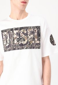 White cotton T-shirt featuring a large, multicolored camouflage logo in dark green, brown, and black, with a circular accent on the sleeve.