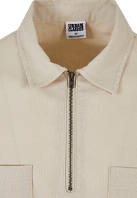 Beige színű, galléros, cipzáros ing, fémből készült cipzárral, két elülső zsebbel és "Urban Classics" felirattal a bélésben.