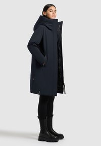 Mørkeblå pufferjakke med høj krave, lynlås foran, sidelommer og en glat overflade. Sammen med sorte knæhøje støvler og tætsiddende strømpebukser.