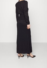 Robe noire à manches longues avec un motif texturé, dotée d'un dos ouvert et d'une taille cintrée. Associée à des chaussures à talons hauts noires.