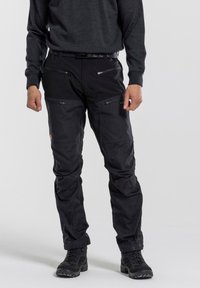 Black outdoor pants made of durable fabric, featuring multiple zipped pockets, a textured surface, and reinforced knee areas. Worn with dark footwear.