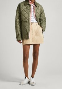 Chaqueta acolchada verde oliva, camisa a rayas, falda beige con bolsillos y zapatillas blancas, mostrando texturas en capas y colores contrastantes.