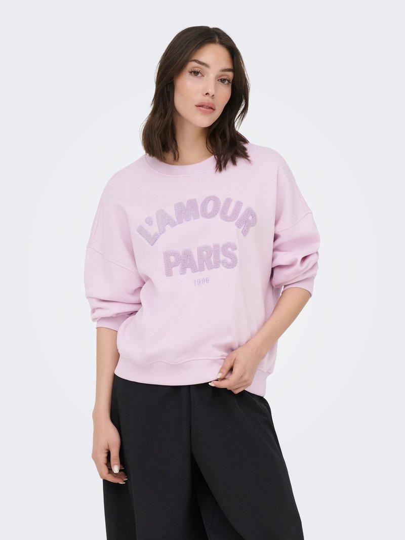Jeune femme aux cheveux châtains mi-longs portant un sweat-shirt rose clair avec le texte "L'AMOUR PARIS 1996" et un pantalon noir à jambes larges.