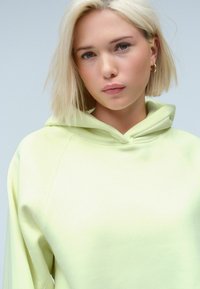 Sweat à capuche vert lime avec une texture douce, présentant une grande capuche et des manches raglan. Le tissu semble lisse et décontracté dans son style.