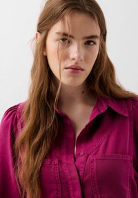 Camicia magenta con bottoni, due tasche sul petto, colletto strutturato e spalle arricciate. Tessuto liscio e vestibilità sartoriale.