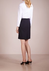 Zwarte pennenrok met een split aan de achterkant, gecombineerd met een wit overhemd met knopen. Het model draagt zwarte open schoenen met hak. Effen achtergrond.