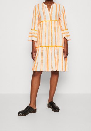 Frau trägt ein knielanges weißes Kleid mit orangen vertikalen Streifen und ausgestellten Ärmeln, kombiniert mit schwarzen Slip-On-Clogs.