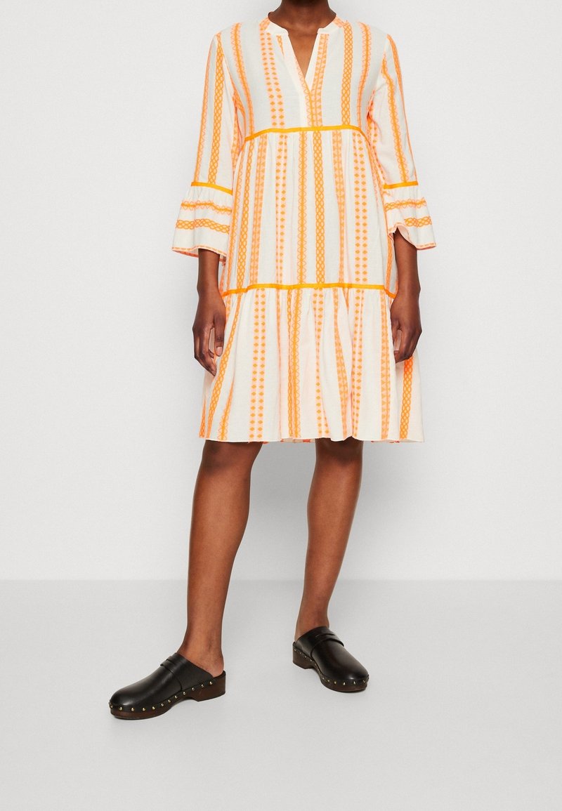 Femme portant une robe blanche mi-longue à rayures verticales orange et manches évasées, associée à des sabots noirs à enfiler.