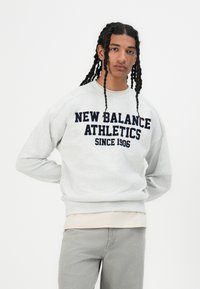 Pelēks kreklveida džemperis ar tumši zilu uzrakstu "NEW BALANCE ATHLETICS SINCE 1906", ar garām piedurknēm un brīvu piegriezumu.