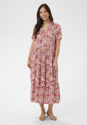 Kobieta uśmiechająca się, stojąca, ubrana w krótką sukienkę o długości do kostek w kwiatowy wzór różowy, czerwony i biały oraz beżowe sandały na obcasie z odkrytymi palcami.