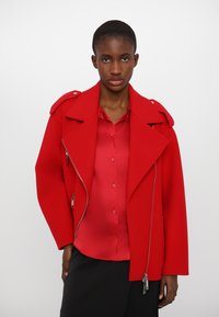 Rote Wollmischjacke mit breitem Kragen, Reißverschluss und Schulterstücken, kombiniert mit einem roten Satinhemd. Strukturierter Stoff und taillierte Passform.