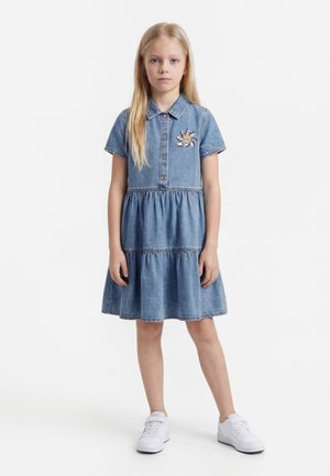 Une fille blonde se tient debout portant une robe en denim bleu à manches courtes avec des boutons, un patch ours de dessin animé, et des baskets blanches sur un fond blanc.