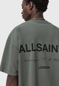 Camiseta de gran tamaño color verde oliva con texto negro en la parte posterior que dice "ALLSAINTS" y "Mensaje para un amigo", con cuello redondo y mangas cortas.