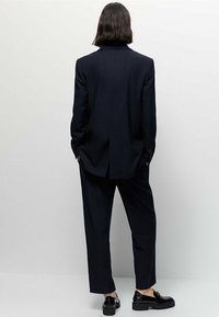 Costume bleu marine foncé avec un blazer ajusté et un pantalon ample, avec un col à cran sur le blazer et des chaussures noires élégantes.