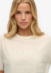 Camiseta de algodón beige con cuello redondo y mangas cortas. Texto decorativo en relieve de un tono contrastante está centrado en el frente.