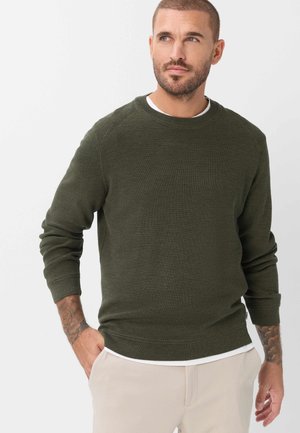 Mann mit kurzen Haaren und Bart, der einen dunkelgrünen Pullover und hellbeige Hose trägt, steht mit einer Hand in der Tasche und schaut zur Seite.