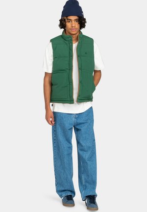 Grøn polstret vest med høj krave, forlommer og brun lynlås. Båret over en hvid t-shirt og parret med løse blå jeans og blå sneakers.