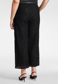 Czarne spodnie z szerokimi nogawkami z koronki, zdobione misternymi kwiatowymi wzorami, z wysokim elastycznym paskiem w talii i falowanym wykończeniem na dole. Noszone z czarnymi sandałami na obcasie.