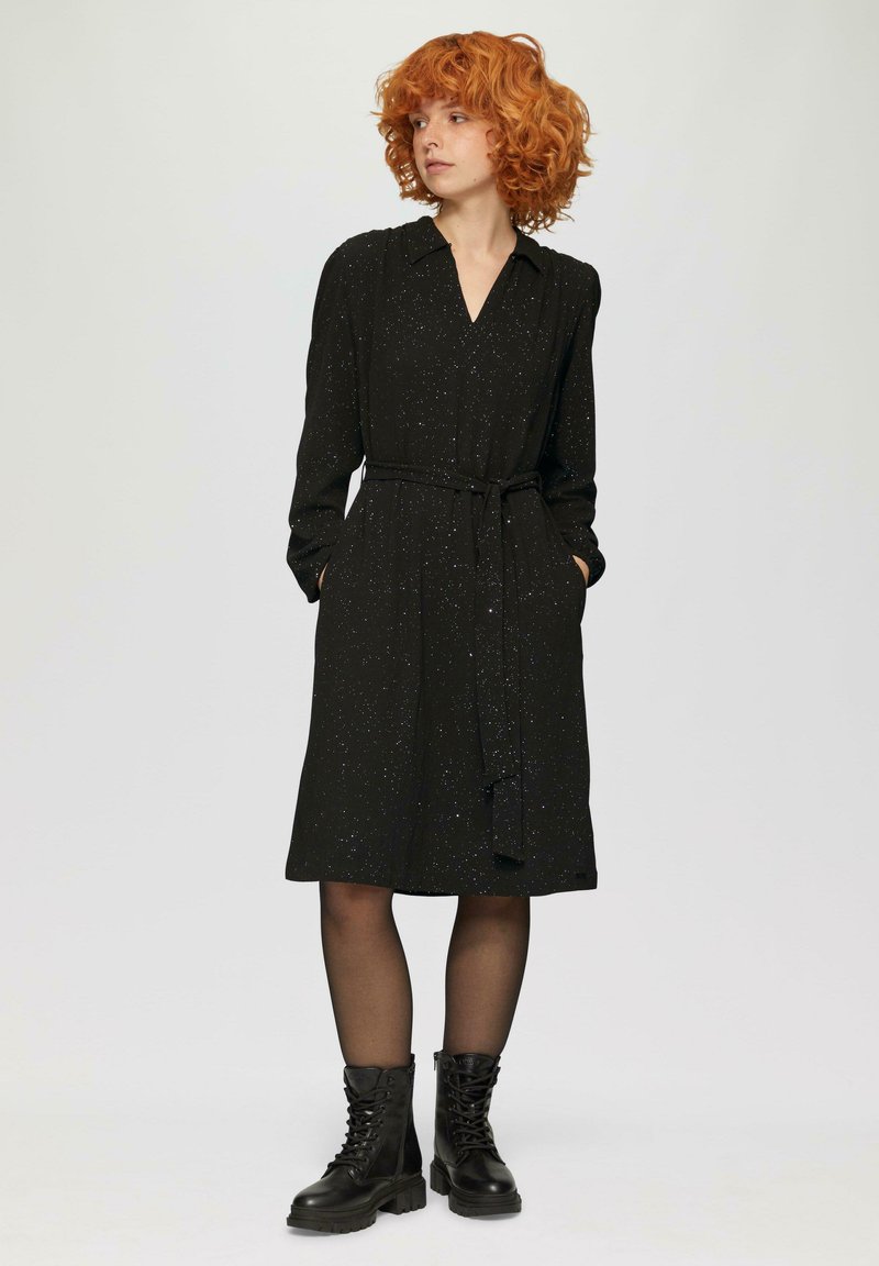 Schwarzes Kleid mit langen Ärmeln, V-Ausschnitt und Gürtel-Detail. Der Stoff hat subtilen Glitzer. Kombiniert mit schwarzen Schnürstiefeln.