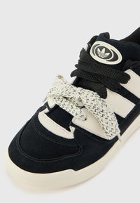 Black suede sneaker with white striped accents, textured black laces, a round logo, and a rubber sole. Features stitched details and reinforced toe.