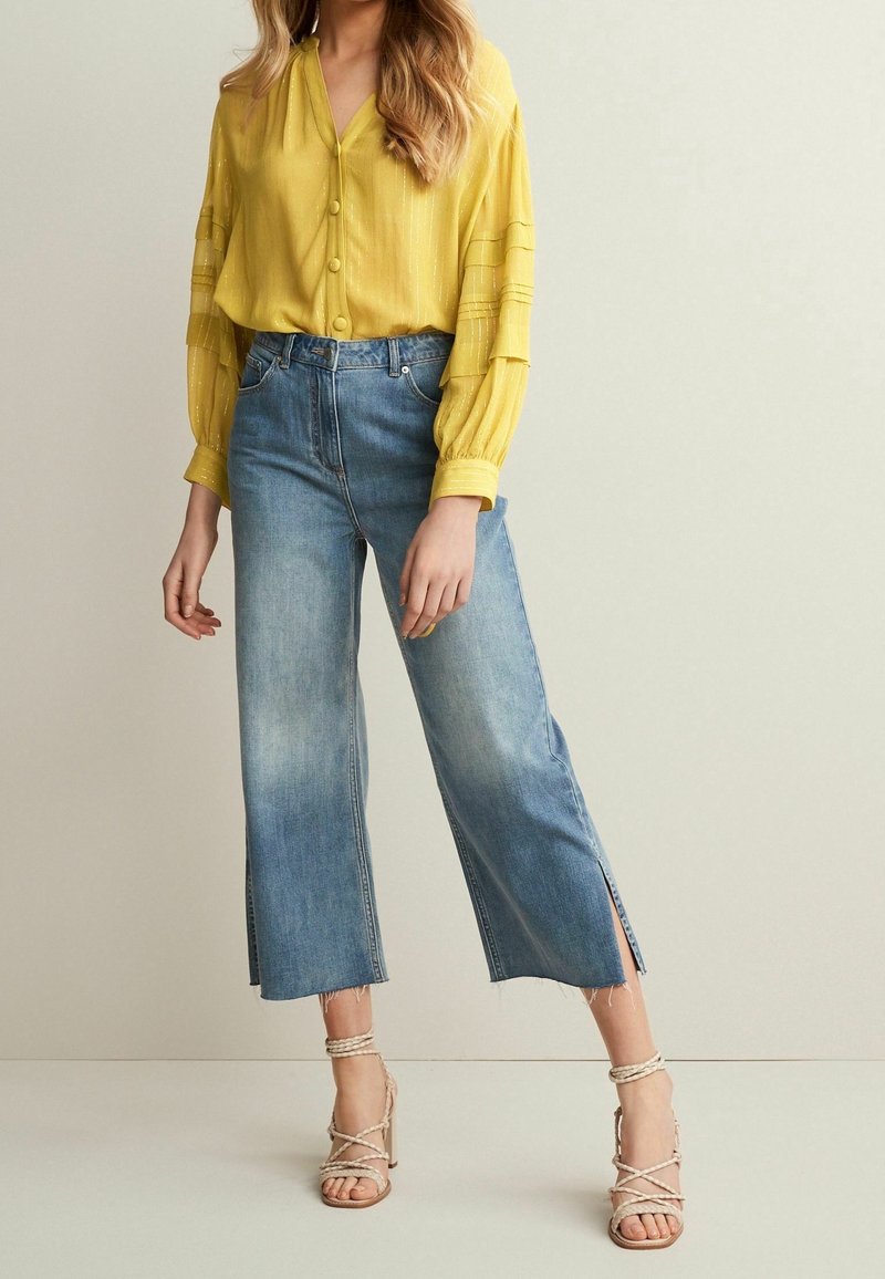 Femme portant un chemisier jaune à manches longues, un jean cropé taille haute à jambes larges avec des fentes sur les côtés, et des sandales à talons hauts beige à brides.