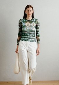 Maglietta a maniche lunghe con un vivace motivo geometrico in verde, blu e rosa, abbinata a pantaloni larghi bianchi e scarpe da ginnastica beige.