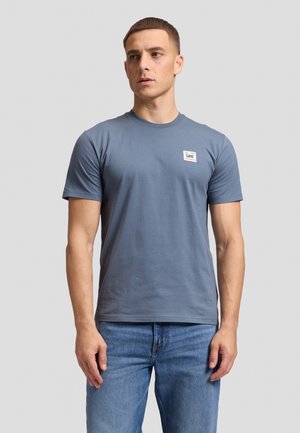 Man wearing a blue short-sleeve t-shirt with a small white Lee logo on the left chest and blue jeans, standing against a plain background.