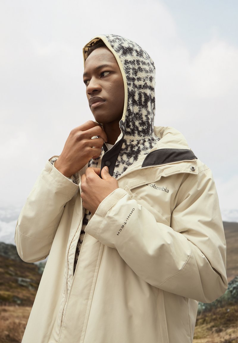 Joven ajustándose una chaqueta beige de Columbia sobre un forro polar con capucha estampado, al aire libre con un cielo nublado y un paisaje montañoso de fondo.