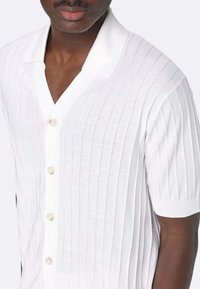 Camicia bianca a maniche corte con una texture a coste, scollo a V e cinque bottoni a contrasto lungo la parte anteriore. Tessuto liscio con una leggera lucentezza.
