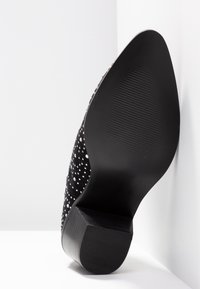 Chaussure noire à bout pointu avec talon bas bloc et dessus clouté, vue par la semelle inférieure sur fond blanc.