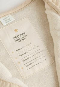 Cremefarbenes Gewebe mit weicher Textur. Ein Etikett zeigt den Text "NEXT baby", einen Stern und Platz für Namen, alles auf einem beigen Hintergrund.
