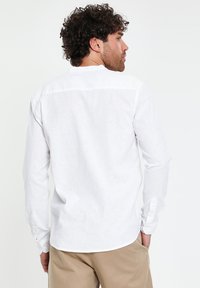 Mann mit lockigem Haar, der ein weißes Langarmshirt und beige Hosen trägt, mit den Händen in den Taschen steht und von der Kamera wegschaut.