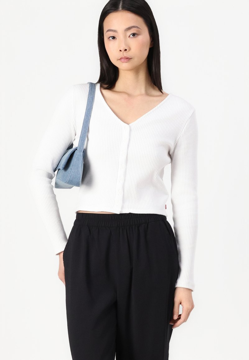 Weißer gerippter Cardigan mit V-Ausschnitt, Knopfverschluss und langen Ärmeln, kombiniert mit schwarzen Hosen mit elastischem Bund und einer blauen Tasche.