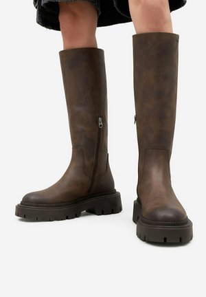 Stivali alti in pelle marrone con suole nere spesse e cerniere laterali, indossati da una persona con gambe nude visibili e gonna nera.