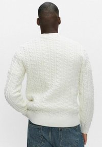 Mies seisoo selin kameraan, päällään valkoinen kaapelineulepaita ja siniset farkut, yksinkertaista vaaleaa taustaa vasten.