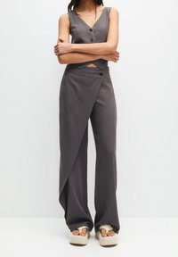 Mujer vestida con un chaleco gris sin mangas y abotonado, pantalones de pierna ancha en un estilo envolvente a juego, y sandalias de plataforma blancas, de pie con los brazos cruzados.