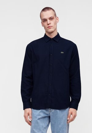 Uomo che indossa una camicia blu navy con bottoni con un piccolo logo verde sul petto e jeans azzurri su uno sfondo semplice.