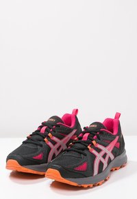 Baskets de course noires avec des accents roses et argentés, tige en tissu texturé, semelle en caoutchouc orange et détails gris sur les côtés.